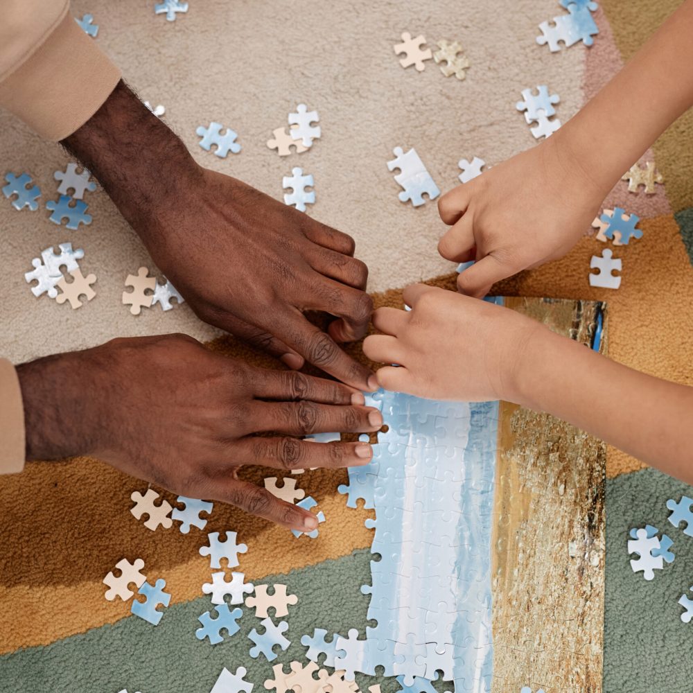 Top view closeup of father and kid solving jigsaw puzzle together on floor at home, focus on hands holding pieces